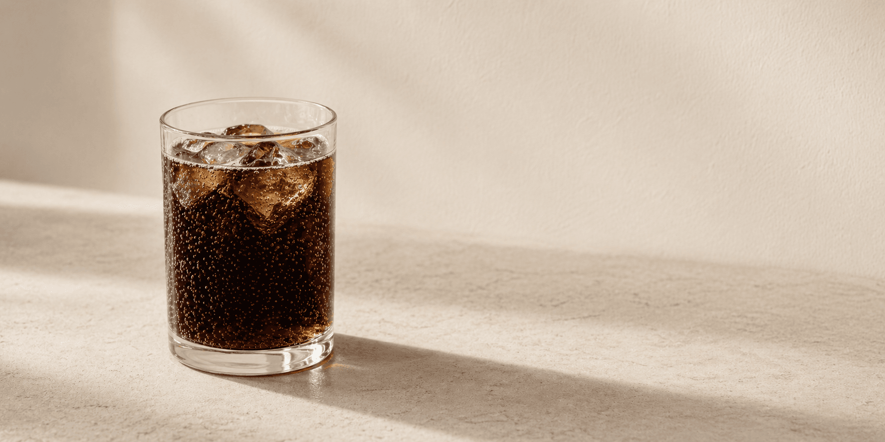 An unbranded glass of dark carbonated diet drink with ice on a warm cream surface.